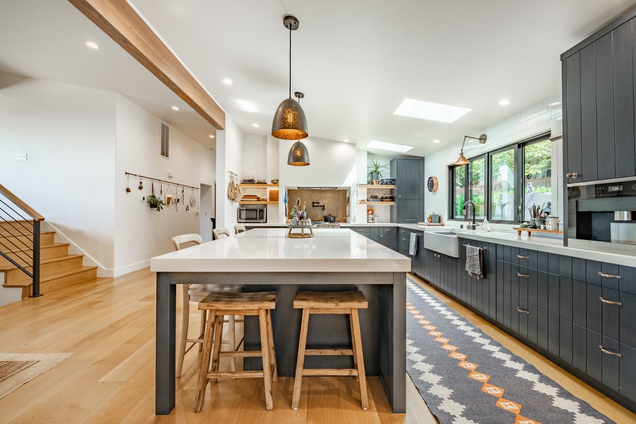 A modern kitchen with dark cabinetry light countertops warm wood flooring. Featuring a large central island with white countertop and seating for four using wooden stools. Two hammered metal pendant lights hang over the island. The interior room is an open concept style with a large window over the sink and a view to the outdoor living space also a view beyond down the entry hallway and family room.